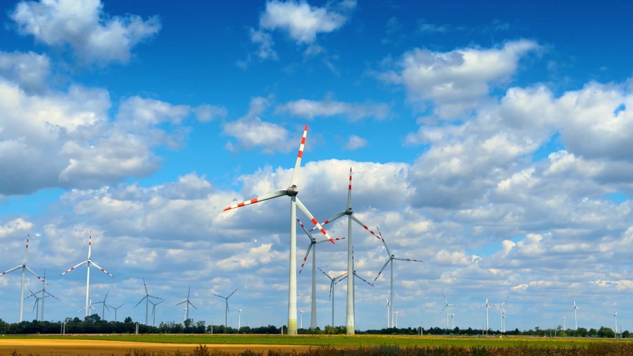 Renewable energy wind farm in open countryside field. White power generators stand on flat agricultural ground under scattered clouds in bright daylight.
