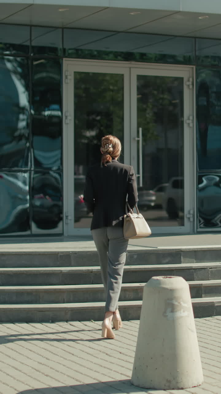 Businesswoman walking towards modern glass office building holding handbag, with sleek reflections of parked cars on facade, showcasing professional lifestyle in urban setting