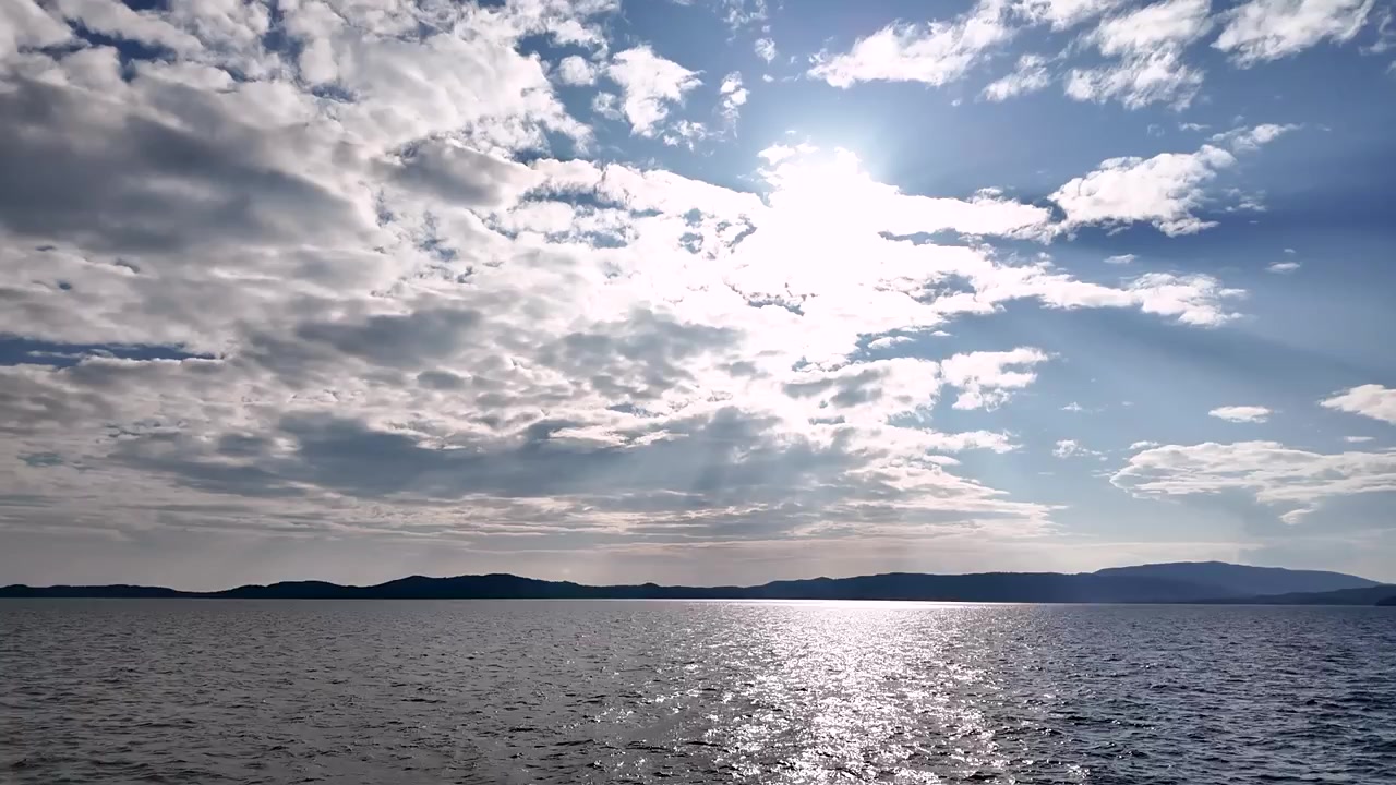 La luce si riflette sull'acqua mentre le nuvole si muovono nel cielo. Il sole splende luminoso, creando un'atmosfera calda sull'acqua. Colline distanti completano la vista.