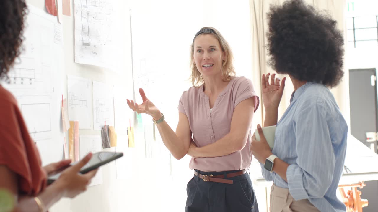 Équipe féminine diversifiée devant un mur, présentatrice en chemisier rose réagissant aux plans épinglés, expliquant la disposition.
