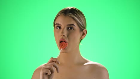 disparo estático medio de una mujer joven y bonita sosteniendo una paleta sabrosa en forma de corazón en la cámara antes de lamerla y disfrutar de la dulzura frente al fondo verde en cámara lenta