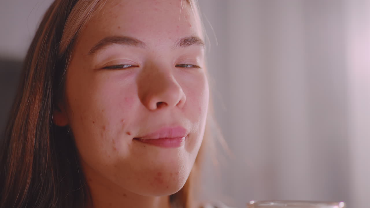 Close up of elegant lady chewing something while holding glass cup, calm indoor background with soft light enhancing natural skin texture and subtle expression