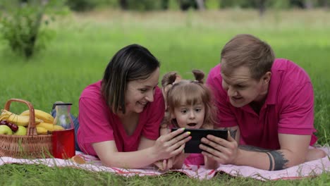 Family-weekend-picnic.-Daughter-child-girl-with-mother-and-father-play-online-games-on-tablet