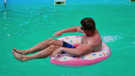 happy caucasian man swimming in the pool. muscular male sits in the inflatable donut rowing with his hands.