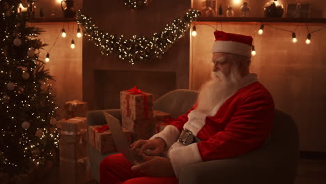 santa claus se sienta junto a un árbol de navidad en un cómodo sofá suave sostiene una computadora portátil en su regazo y responde correos electrónicos de niños pequeños.