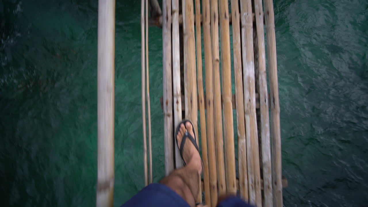 Vista in prima persona mentre si cammina su uno stretto ponte di bambù sopra un mare turchese e limpido a Carabao Island, vicino a Boracay, Filippine, con gambe e sandali visibili che si avvicinano alla riva rocciosa.