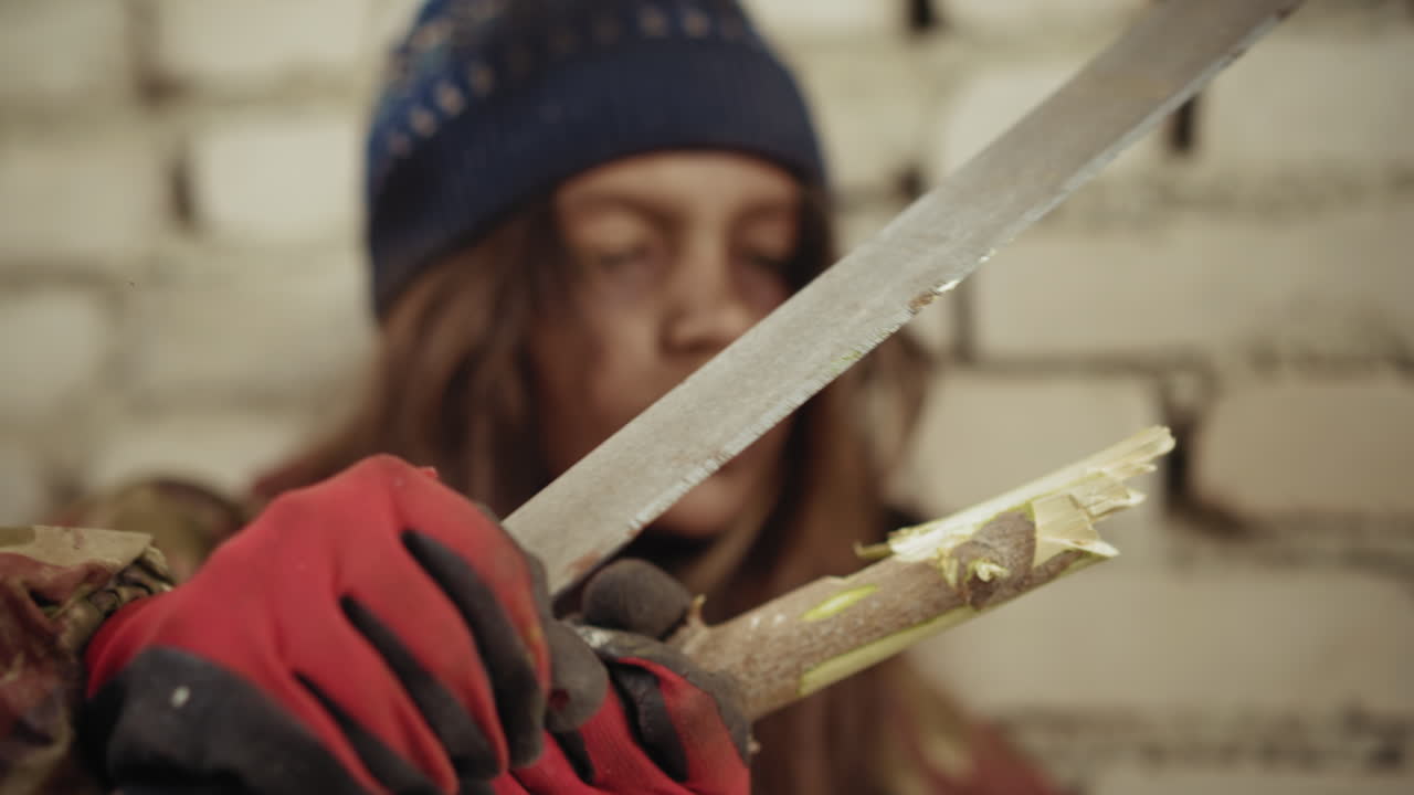 Close up of young lady wearing gloves and winter hat carving stick with knife in rugged outdoor environment, demonstrating survival skill and focus, set against damaged brick wall