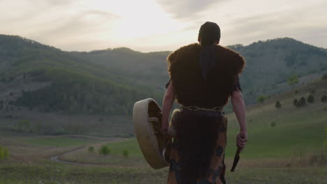 actor walks carrying shamanic drum against old hills