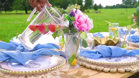 Fruity-water-with-strawberries-is-being-poured-into-a-glass-with-mint-lemon-and-cucumber-and-straw-inside-outdoor-picnic-children-party-in-nature-slow-motion