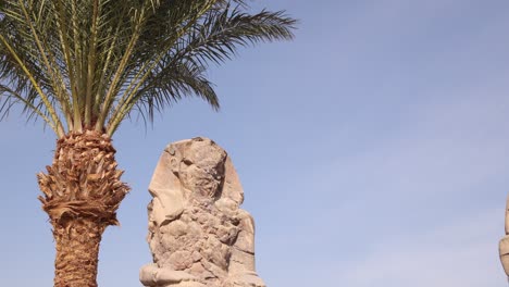 Ruinen-Der-Riesigen-Statue-Des-Pharaos-Mit-Bergen-Im-Hintergrund-Bei-Den-Kolosse-Von-Memnon-In-Luxor,-Ägypten-Mit-Palme