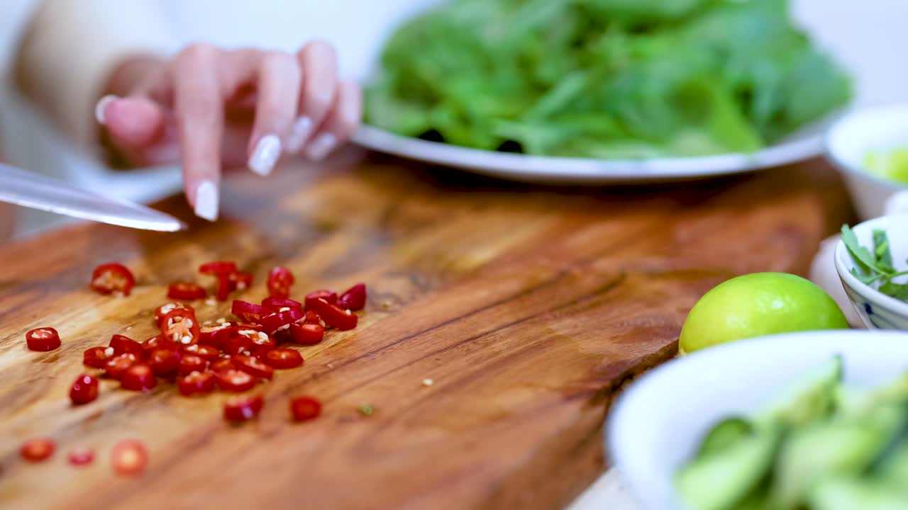 A person slices red chilies on a wooden board, surrounded by fresh ingredients, under bright lighting