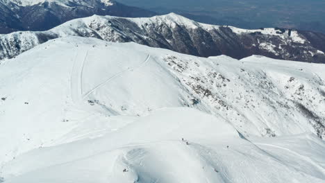 Skiers-dot-a-vast-snowy-mountainscape-under-a-blue-sky,-serene-winter-sports-scene,-aerial-view