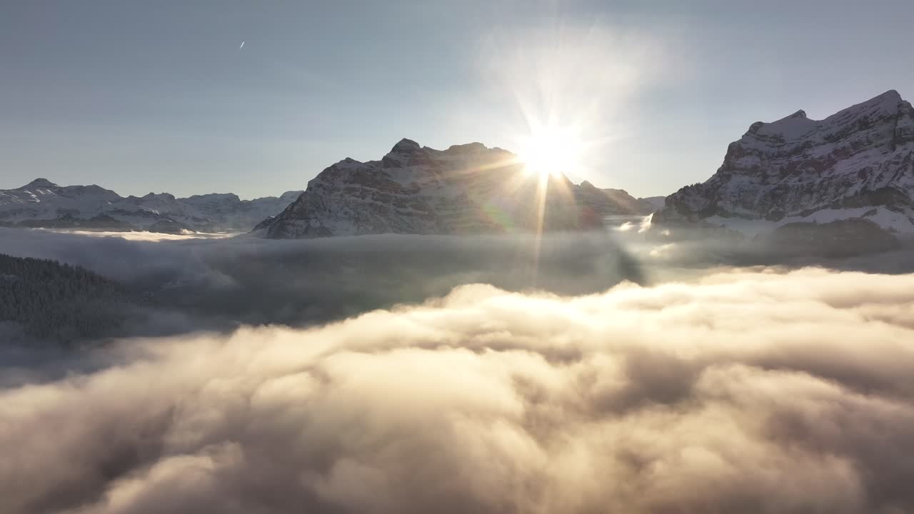 Drone flight above the clouds with the sun backlighting the Glärnisch mountain range in the Glarus Alps, Switzerland.