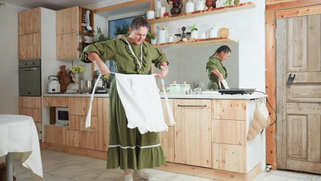chef in green gown picks up white apron and begins tying it around waist while standing barefoot in cozy rustic kitchen filled with wooden cabinets, modern appliances, and reflected mirror view