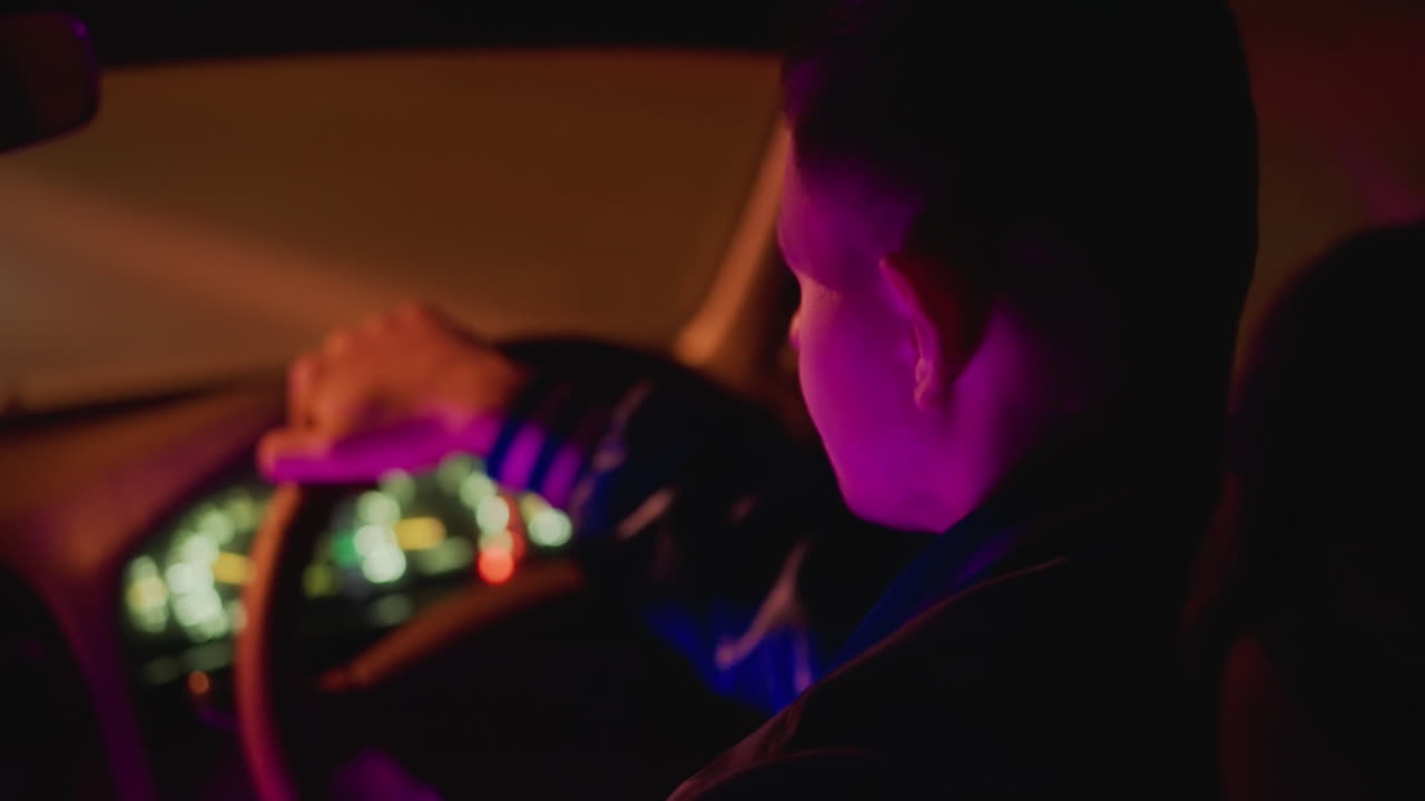 Bilinteriör nattetid med vit man som tittar på neonupplyst instrumentbräda, händer på ratten, långsam stadskörning, lila och rosa sken, filmisk kornighet och tät bildutsnitt som förmedlar ensamhet och eftertanke