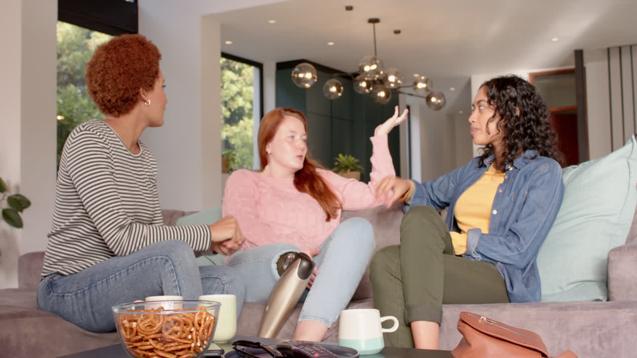 Chatting and relaxing, diverse female friends sitting on couch with snacks and drinks