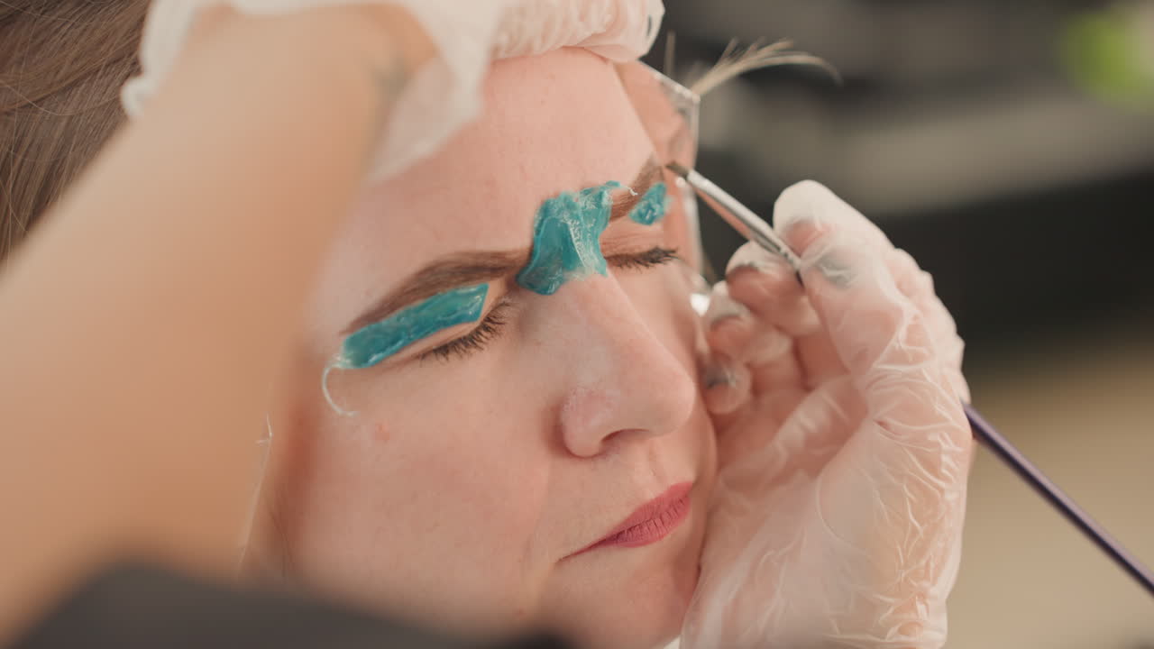 White woman receiving eyebrow tinting and foil highlight in salon, aesthetician applies teal pigment with brush and gloves, closeup showing precise shaping and sterile technique, relaxed client