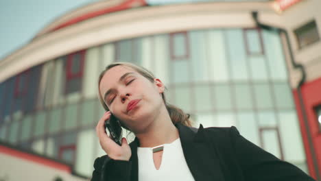 Furious lady in professional outfit speaks on phone while visibly tired and upset, standing in urban setting with people blur in distant background and tall modern glass building behind her