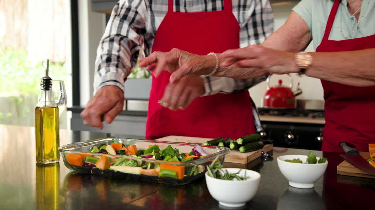 Senior couple in kitchen preparing festive meal, pouring oil on vegetables, at home