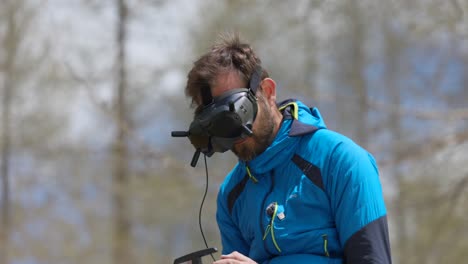 man in blue jacket using a virtual reality headset and remote control to navigate a drone in an outdoor setting