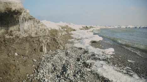 Frozen-Shoreline-With-Icicles-On-Rocks-In-Splashing-Ocean-Waves-At-Sunny-Winter-Day