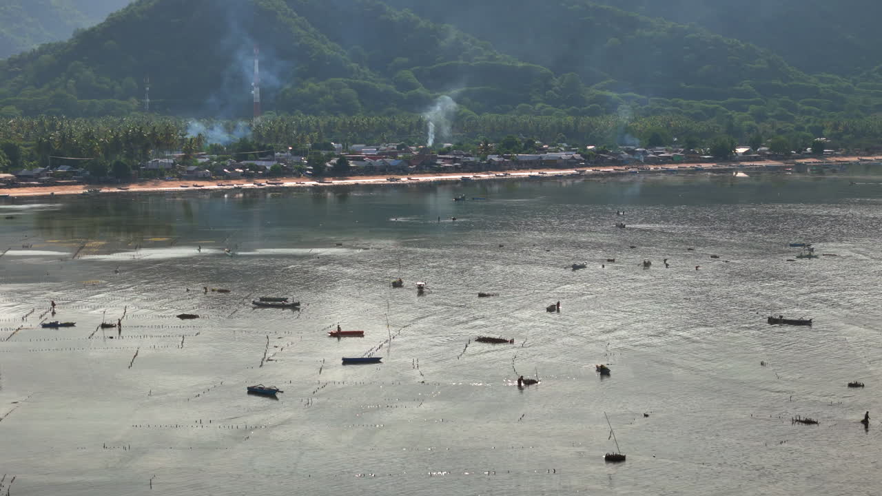 ภาพมุมสูงจากโดรนของหมู่บ้านชาวประมงชายฝั่งในเวลาน้ำลง พร้อมเรือเล็กลอยอยู่ในน้ำตื้นและเนินเขตร้อนสีเขียว แนวชายฝั่งโค้งไปตามอ่าว โดยมีต้นปาล์มและเนินเขียวชอุ่มหนาทึบตั้งตระหง่านอยู่ด้านหลัง
