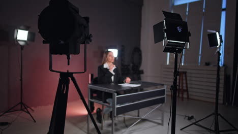 professional illumination equipment is set in studio around a woman sitting at desk. blogger talks to camera and gestures with her hands.