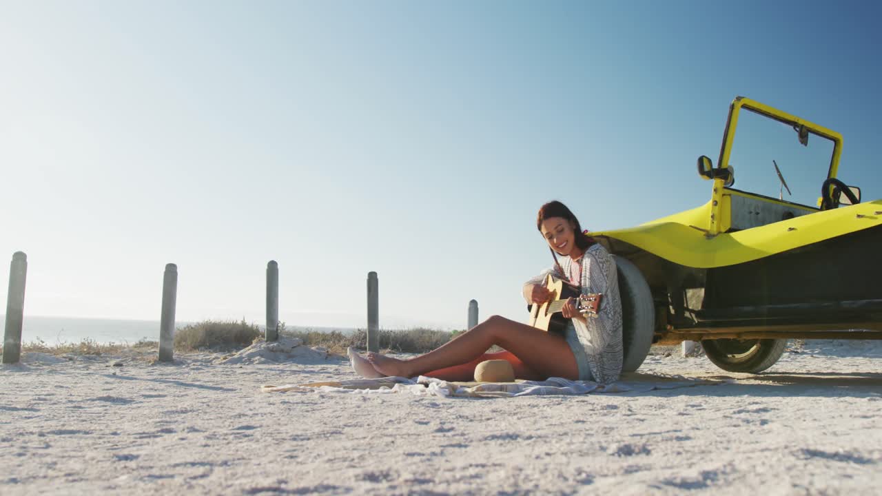 Glad kaukasisk kvinde sidder ved siden af en strandbuggy ved havet og spiller guitar.