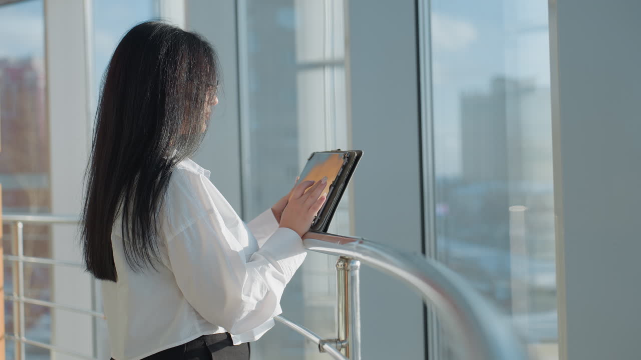 Side view of investor in white blouse standing at iron railing using tablet, with screen reflection visible on glass window, surrounded by modern architecture and bright natural daylight