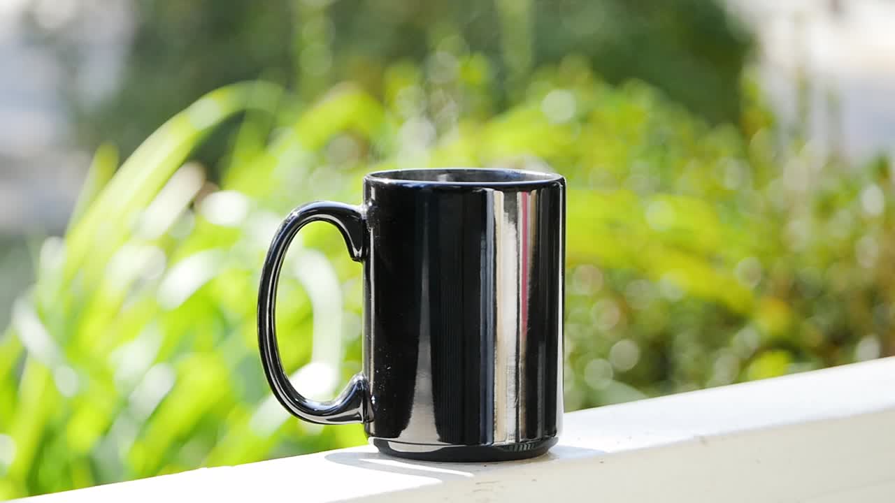 suave toma en cámara lenta de una taza negra llena de café caliente sentada y humeante en un porche delantero de atlanta con un soleado bokeh de arbusto que simplifica el fondo