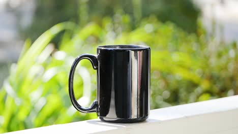 Smooth-slow-motion-shot-of-a-black-mug-full-of-hot-coffee-sitting-and-steaming-on-an-Atlanta-front-porch-with-sunny-bush-bokeh-making-the-background-simple