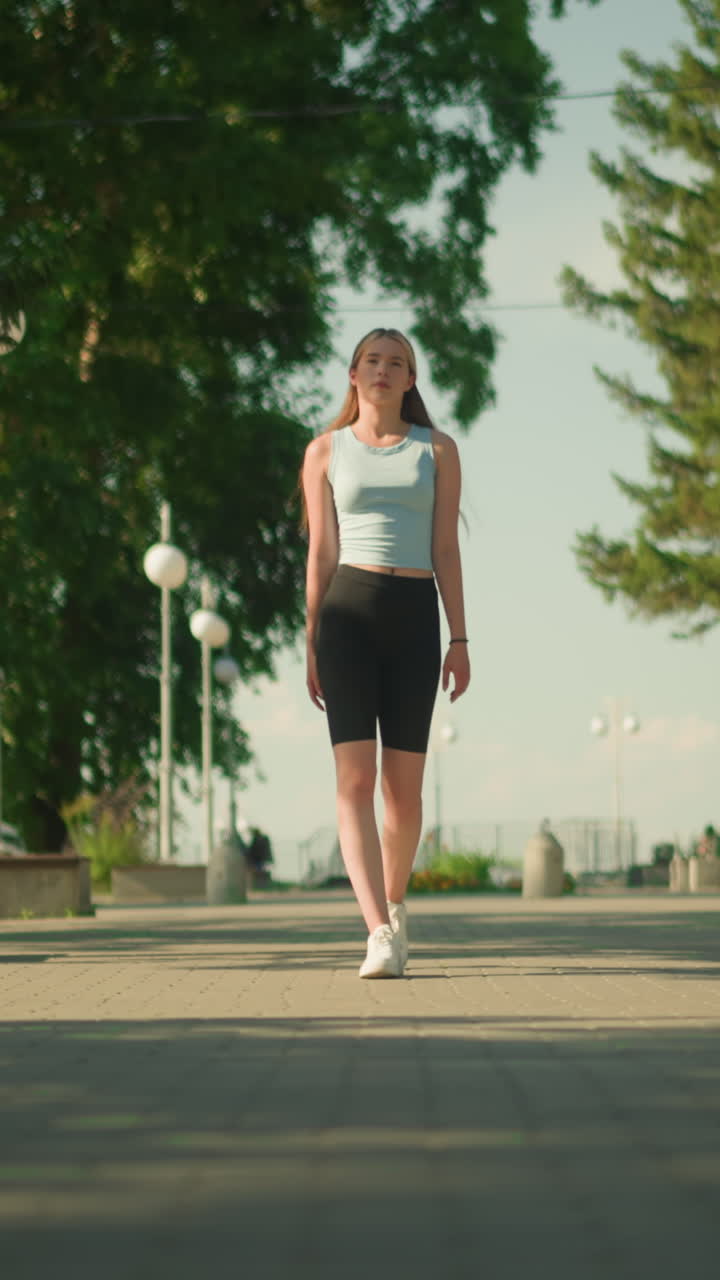 dama caminando con confianza en el camino entrelazado alineado con postes de luz, rodeado de árboles altos, vegetación exuberante, coche estacionado, y vista borrosa de persona en la distancia empujando cochecito de niño