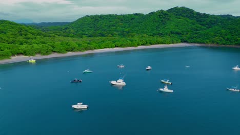 Circling-shot-of-fishing-boats-anchored-near-the-shore-in-deep-blue-waters-with-the-forest-and-sky-in-the-background