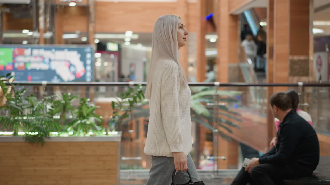 urban woman holding black bag walking through modern mall walkway admiring crystal decor under bright ceiling lights near seating corner capturing casual style and curious expression