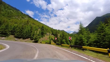 car driving on a mountain, forest road, surrounded by tall and green trees with a clear blue sky and white clouds, transfagarasan, romania