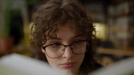 Primer-Plano-De-Una-Estudiante-Segura-De-Sí-Misma,-Con-Cabello-Rizado-Y-Gafas,-Leyendo-Un-Libro-En-La-Biblioteca.