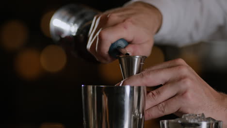 Primer-Plano-De-Un-Barman-Midiendo-Licor-En-Un-Jigger-Y-Vertiéndolo-En-Una-Coctelera-De-Metal.