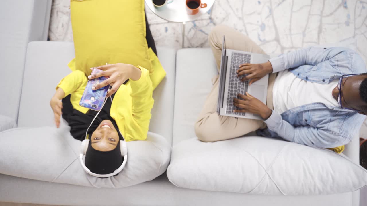 una alegre y feliz pareja de casados africanos sentados juntos en el sofá y ocupados con aparatos tecnológicos.