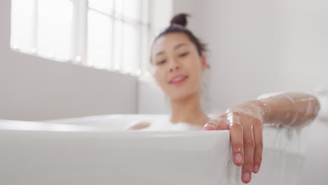 Video-of-portrait-of-smiling-biracial-woman-sitting-in-bathtub-in-bathroom