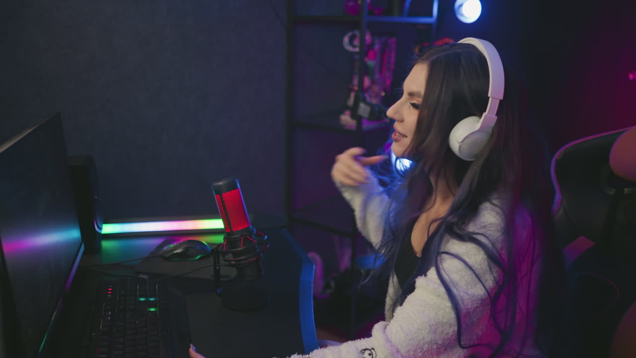 happy young woman wearing headphones and cozy sweater leans forward smiling while adjusting position at computer desk after winning game, surrounded by glowing lights and microphone in modern studio