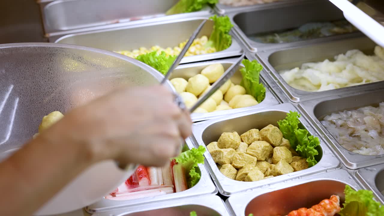 Person uses tongs to pick hotpot ingredients at brightly lit Asian buffet self-service counter