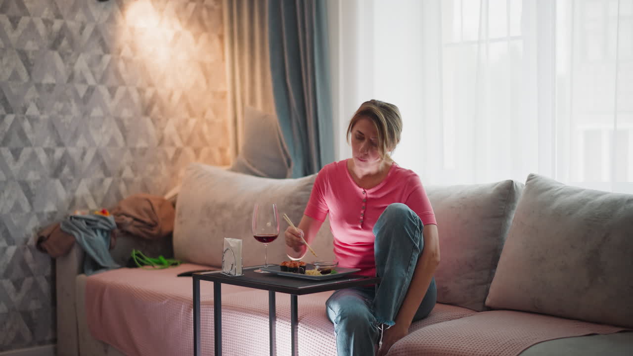 Woman seated on sofa enjoying meal with chopsticks and wine beside her in relaxed home setting as light filters through curtain while casual clothes are placed at end of couch