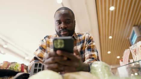 Shooting-from-below-a-man-with-Black-skin-color-walks-around-the-supermarket-with-a-wheelbarrow-and-looks-at-a-black-phone,-where-a-list-of-necessary-products-is-written