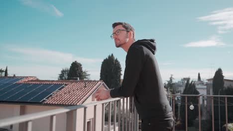 young attractive caucasian man with beard and glasses on balcony in sunshine watching surrounding, enjoying good weather sunshine