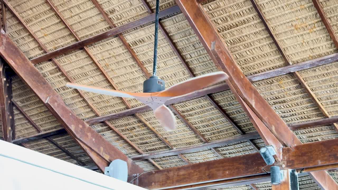 A wooden ceiling fan spins under a straw roof in a tropical setting, with natural lighting highlighting the rustic architecture