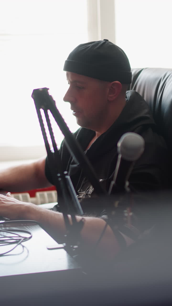 음악 프로듀서 또는 드캐스터인 한 남자가 스튜디오 방에서 컴퓨터 앞에 앉아 검은 옷과 모자를 입고 있습니다. 스튜디오에는 마이크와 비언트 조명이 장착되어 있습니다.