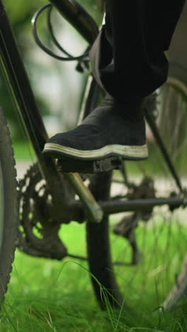 primer plano de alguien colocando el pie en el pedal de una bicicleta estacionada apoyándose en un árbol en un campo cubierto de hierba, centrándose en la bicicleta y los componentes, comprobando la posición, con un fondo verde borroso