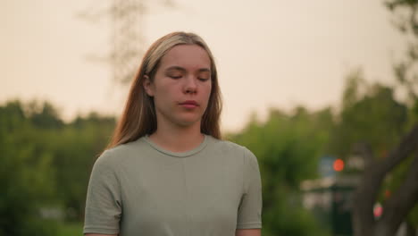 close-up of young woman with tired, contemplative expression walking outdoors, dressed in a casual green shirt, set against a natural, blurred background of greenery in soft light