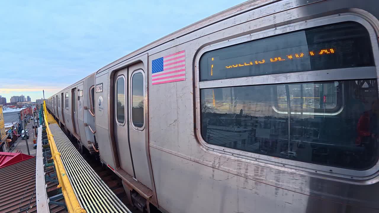 New York, USA, 14. marts 2026: New York City metrotog på hævede spor i skumringen. Sølvfarvet metrovogn med amerikansk flag-klistermærke bevæger sig langs en hævet jernbanelinje med byens skyline i baggrunden.