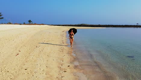 Attractive-young-woman-with-beach-umbrella-walking-on-sand-washed-by-clear-sea-water-on-a-bright-blue-sky-background-in-Bali